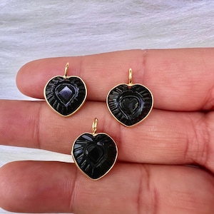 Könnte beinhalten: Drei schwarze herzförmige Anhänger mit goldfarbenen Ösen. Jeder Anhänger hat ein geschnitztes Herzdesign in einem herzförmigen Rahmen. Die Anhänger werden vor einem weichen, weißen Hintergrund präsentiert.