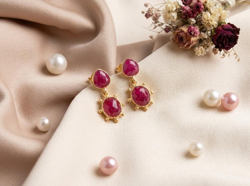 May include: A pair of gold drop earrings featuring two faceted, deep pink ruby-like stones each. The top stone is pear-shaped, while the bottom stone is oval with a sunburst-style gold setting. The earrings are displayed alongside scattered pearls.