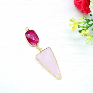 May include: A gold-toned pendant with a rectangular, faceted, magenta-colored gemstone at the top and a light pink, triangular gemstone at the bottom. The gemstones are connected by a gold chain. The pendant is set against a white background with flowers.