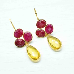 May include: A pair of gold-toned earrings featuring a cluster of three oval, deep red gemstones and a single, teardrop-shaped yellow gemstone. The earrings have a hook closure and are set against a white background.