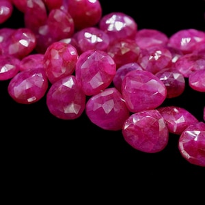 May include: Close-up of a collection of faceted, heart-shaped ruby gemstones. The gems are a deep, vibrant pinkish-red colour, with multiple facets catching the light. The background is solid black, highlighting the gems.