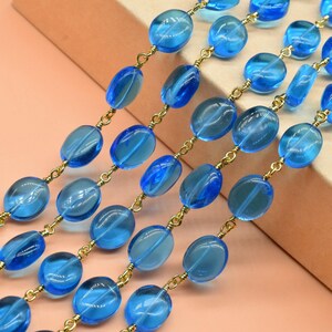 May include: Close-up of multiple strands of blue glass bead necklaces. The beads are oval-shaped and strung on gold-colored chains. The beads vary in shades of blue, creating a gradient effect. The background is a light pink and brown.