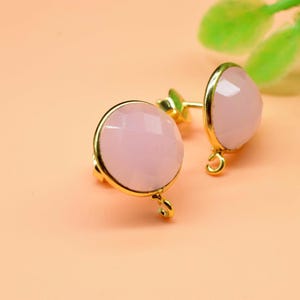 Puede incluir: Un par de pendientes de botón dorados con gemas facetadas de color rosa pálido. Los pendientes tienen forma redonda y un pequeño lazo en la parte inferior. Fondo melocotón suave con un toque de follaje verde.