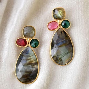 Puede incluir: Un par de pendientes dorados con piedras de labradorita en forma de lágrima. Cada pendiente tiene un grupo de piedras preciosas más pequeñas en tonos rojos, verdes y gris verdoso. Las piedras tienen una superficie facetada.