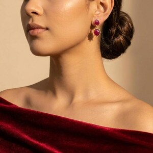 May include: A pair of gold drop earrings featuring two faceted, deep pinkish-red oval gemstones. The top stone is smaller and rounder, while the bottom stone is larger and surrounded by a sunburst-style metal frame. The earrings are worn with a dark red velvet off-the-shoulder garment.