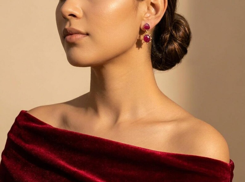 May include: A pair of gold drop earrings featuring two faceted, deep pinkish-red oval gemstones. The top stone is smaller and rounder, while the bottom stone is larger and surrounded by a sunburst-style metal frame. The earrings are worn with a dark red velvet off-the-shoulder garment.