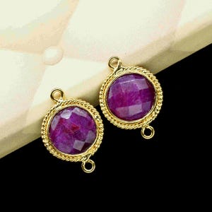 May include: Two round, gold-colored jewelry components with a faceted, deep magenta gemstone center. The gold setting has a twisted rope design around the gemstone. Each component has a small loop at the top and bottom.