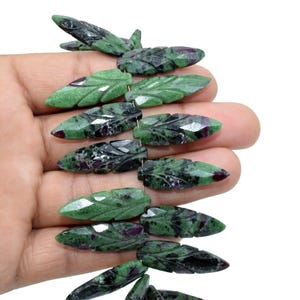 Peut inclure: Un fil de perles de pierres précieuses sculptées en forme de feuille. Les perles sont vertes avec des inclusions vert foncé et rouges. Les perles sont tenues dans une main sur fond blanc. Les perles mesurent environ 2,5 cm de long.