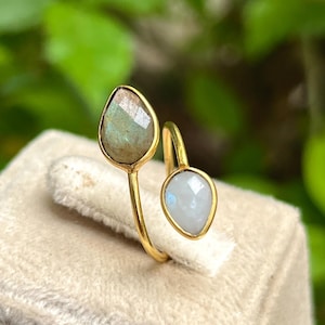 Könnte beinhalten: Ein goldfarbener Ring mit zwei tropfenförmigen Edelsteinen. Einer ist ein gedämpftes Grün, der andere ein blasses Blau. Der Ring hat ein offenes Design, die Edelsteine sind in Gold gefasst.