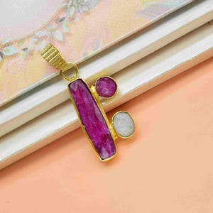 Puede incluir: Un colgante dorado con una gema rectangular magenta grande, acentuada por dos gemas redondas más pequeñas. El colgante se encuentra sobre un fondo melocotón, lo que sugiere un tema de joyería o accesorios.