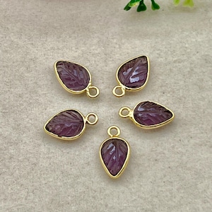 May include: Five leaf-shaped pendants with a carved leaf design. The pendants are a deep purple color with gold-colored metal edging and a small loop at the top for attaching to a chain or other jewelry. The pendants are arranged on a light gray surface.
