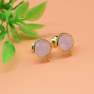 May include: A pair of stud earrings with light pink, faceted gemstones set in gold-colored metal. The earrings are displayed on a peach-colored surface, with a sprig of green leaves in the background. These earrings are a stylish accessory.