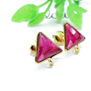 Pode incluir: Brincos de botão dourados com pedras preciosas triangulares, facetadas, vermelho rubi. Os brincos são cravejados em uma moldura dourada e possuem fecho de pressão. Fundo branco com folhas verdes.