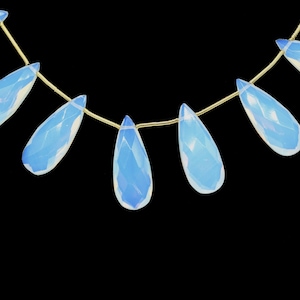 May include: Strand of teardrop-shaped, faceted opalite beads. Each bead is a pale blue with a pearlescent sheen, strung on a thin, gold-colored cord. The beads are displayed against a black background.