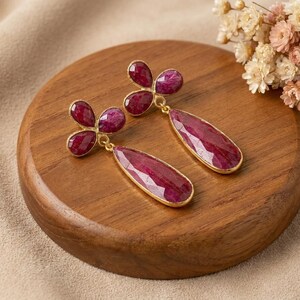 May include: A pair of gold-toned drop earrings featuring faceted, deep magenta-red gemstone clusters. Each earring consists of a three-petaled floral top connected to a long, teardrop-shaped pendant, all set in a thin gold-colored metal frame.