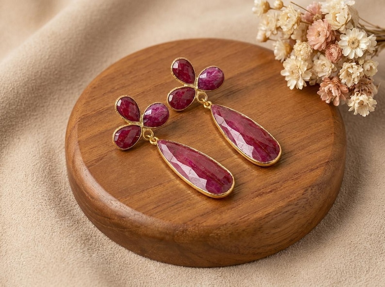 May include: A pair of gold-toned drop earrings featuring faceted, deep magenta-red gemstone clusters. Each earring consists of a three-petaled floral top connected to a long, teardrop-shaped pendant, all set in a thin gold-colored metal frame.