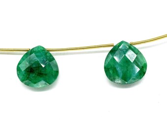 Op de afbeelding: Twee gefacetteerde, peervormige smaragd edelstenen geregen aan een dunne gouden draad. De smaragden hebben een levendige groene kleur met zichtbare interne patronen. De achtergrond is effen wit, waardoor de edelstenen worden benadrukt.