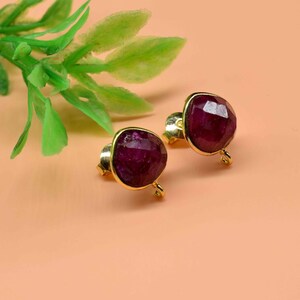 Pode incluir: Um par de brincos de rubi com engastes dourados. Os brincos apresentam pedras preciosas facetadas de cor vermelha escura. O fundo é de cor pêssego sólida, com um raminho de folhas verdes no canto superior esquerdo.