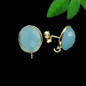 May include: A pair of gold-coloured stud earrings featuring oval, light blue gemstones. The gemstones have a faceted cut and are set in a gold bezel. The earrings are shown against a black background.