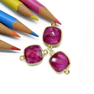 Könnte beinhalten: Drei quadratische, magentafarbene Edelsteinanhänger mit goldfarbenen Fassungen. Die Anhänger befinden sich in der Nähe einer Sammlung von Buntstiften, darunter rot, orange, blau, gelb und rosa.