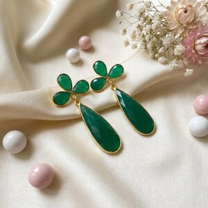 May include: A pair of gold-tone drop earrings featuring faceted dark green teardrop-shaped stones suspended from a three-leaf clover design. Each earring is crafted with a polished gold metal setting that frames the vibrant green gemstone elements.