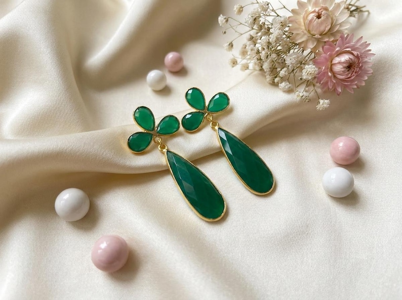 May include: A pair of gold-tone drop earrings featuring faceted dark green teardrop-shaped stones suspended from a three-leaf clover design. Each earring is crafted with a polished gold metal setting that frames the vibrant green gemstone elements.