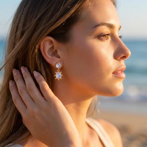 May include: A gold drop earring featuring a small round moonstone stud connected to a larger oval moonstone pendant. The oval pendant is framed by a gold border with small radiating spikes, creating a sunburst effect. The stones have a soft blue glow.