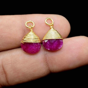 May include: Two gold-colored, wire-wrapped pendants with teardrop-shaped, faceted, magenta-colored gemstones. Each pendant has a small loop at the top for hanging. The background is black.