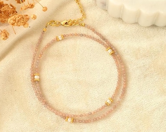 Könnte beinhalten: Eine zarte Halskette mit kleinen, hellrosa Perlen und goldfarbenen Akzenten. Die Halskette hat ein doppelreihiges Design mit kleinen perlenartigen Verzierungen. Der Verschluss und die Kette sind goldfarben. Der Schmuck wird auf einem cremefarbenen Stoff präsentiert.