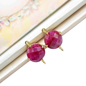 Puede incluir: Dos cuentas de piedras preciosas redondas y facetadas, de color rojo rubí, con conectores dorados. Las cuentas se muestran sobre una superficie blanca con un diseño floral en el fondo. Las piedras preciosas tienen una superficie pulida y reflectante.