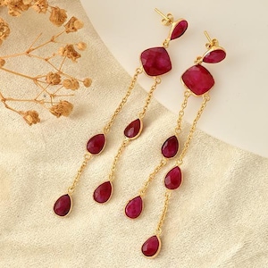 Peut inclure: Boucles d'oreilles dorées avec un design pendant. Chaque boucle d'oreille présente une pierre précieuse rouge foncé taillée en carré en haut, suivie d'une chaîne de petites pierres rouges en forme de goutte. Sur un tissu crème.