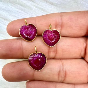 May include: Three heart-shaped pendants with a deep magenta hue and gold-colored settings. Each pendant has a textured surface and a small loop at the top for hanging. The pendants are displayed against a soft, white background.