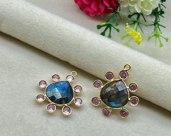 May include: Two gold-toned pendants, each featuring a central, faceted, blue-green gemstone surrounded by small, pink gemstones. The pendants have a floral-like design. The background includes flowers and a rolled fabric.