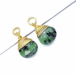 Op de afbeelding: Twee druppelvormige hangers met gefacetteerde groene en zwarte edelstenen, gewikkeld in goudkleurig draad. Elke hanger heeft een kleine lus aan de bovenkant. De achtergrond is wit met een diagonale lijn.