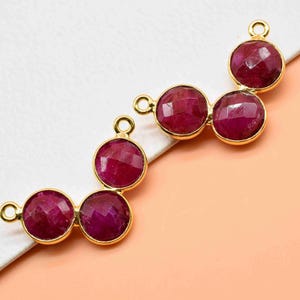 Puede incluir: Dos componentes de joyería dorados, cada uno con tres piedras preciosas redondas, facetadas, de color rojo intenso. Las piedras están engastadas en biseles dorados con pequeños bucles para la fijación. Los componentes están dispuestos sobre un fondo blanco y melocotón.