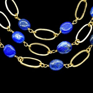 May include: Close-up of three gold necklaces with oval links and deep blue lapis lazuli beads. The necklaces are layered against a black background, showcasing the contrast between the gold and the blue gemstones.