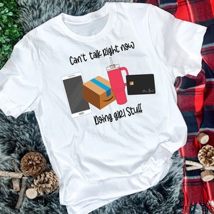 May include: White t-shirt with the text "Can't talk right now doing girl stuff" and illustrations of a smartphone, a box, a pink tumbler, and a credit card.