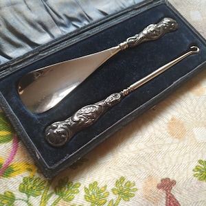 Puede incluir: Un calzador de zapatos de plata vintage y un gancho para cordones de zapatos en una caja forrada de terciopelo negro. Las herramientas tienen diseños ornamentados.