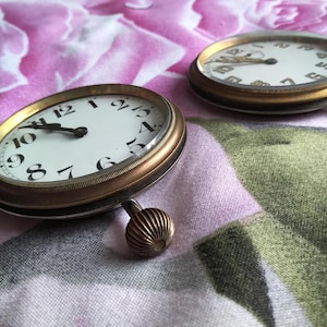 May include: Two antique gold pocket watches with white faces and black numbers on a floral patterned fabric background.