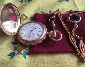 Puede incluir: Un reloj de bolsillo de oro con una esfera blanca y marcadores de hora romanos negros. El reloj está abierto y la parte posterior es visible. El reloj está sobre una bolsa de terciopelo rojo con una cadena de oro.
