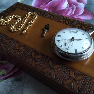May include: A silver pocket watch with a white face and black Roman numerals rests on a wooden box. A gold chain and key are next to the watch. The box has a carved floral design.