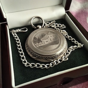 May include: A silver pocket watch with an engraved image of a house and a chain in a black velvet box.