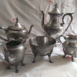 May include: A silver tea set with an ornate design. The set includes a teapot, creamer, sugar bowl, and a serving bowl. All pieces have a silver finish and intricate details.