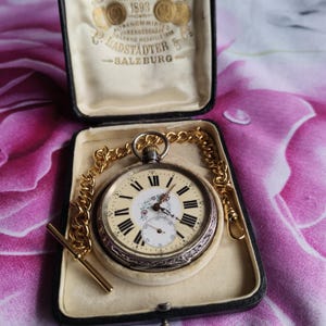 May include: A silver-toned pocket watch with a cream-colored face, black Roman numerals, and a floral center design. It features a gold-toned chain with a T-bar and rests in a black presentation box with text reading C. LADSTÄDTER & CO SALZBURG.