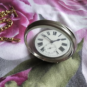 May include: A silver pocket watch with a white face and black Roman numeral hour markers. The watch is in a silver case with a glass cover. The watch is on a pink and white floral fabric.