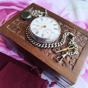 May include: A silver pocket watch with a white face, black Roman numerals, and a sub-dial, attached to a silver chain with a brass winding key. The watch rests on a rectangular wooden box featuring intricate carved floral patterns on its lid.
