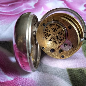 Könnte beinhalten: Eine silberne Taschenuhr mit einem goldfarbenen Zifferblatt und aufwendigen Gravuren. Die Uhr ist geöffnet und zeigt das Innenleben des Mechanismus.