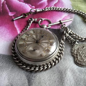 May include: A silver pocket watch with a white face and black Roman numeral hour markers. The watch is attached to a silver chain with a decorative fob.