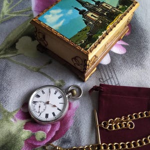 May include: A silver pocket watch with a white face and black Roman numeral hour markers. The watch is attached to a gold chain and is lying on a floral patterned fabric with a wooden box in the background.