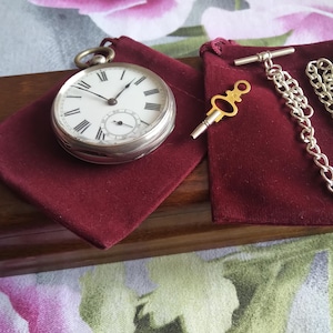 May include: A silver pocket watch with Roman numeral markings on a white face, a silver chain, and a key, all displayed on a burgundy velvet pouch.